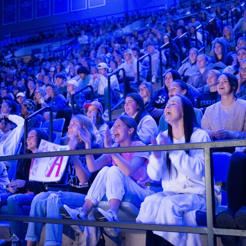 A large crowd of Indiana State University students enthusiastically watches a performance during Entertainment Night, illuminated by blue stage lighting.